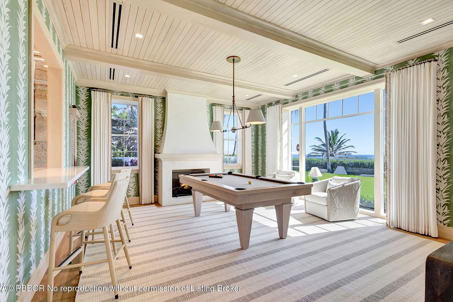 This is a bright and airy living room featuring a pool table as a focal point. The room is decorated with a green and white leaf-patterned wallpaper, complemented by white curtains and trim. Large windows offer a view of the ocean, and the space is furnished with comfortable seating, creating a relaxed and luxurious atmosphere.
