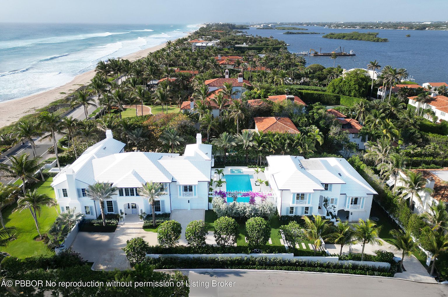 This aerial shot showcases a luxurious beachfront estate with a pristine white exterior, complemented by lush tropical landscaping. A sparkling blue pool sits between the main house and a guest house, while the property extends towards a sandy beach and the ocean. The overall impression is one of opulence and serene coastal living.