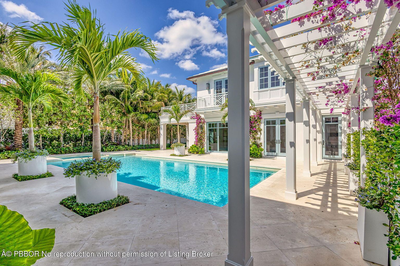 This image showcases a luxurious backyard pool area, featuring a pristine blue pool surrounded by light-colored stone paving. Lush tropical landscaping, including palm trees in decorative planters, enhances the serene atmosphere. A white pergola adorned with flowering vines adds an elegant touch, complementing the white exterior of the house in the background.