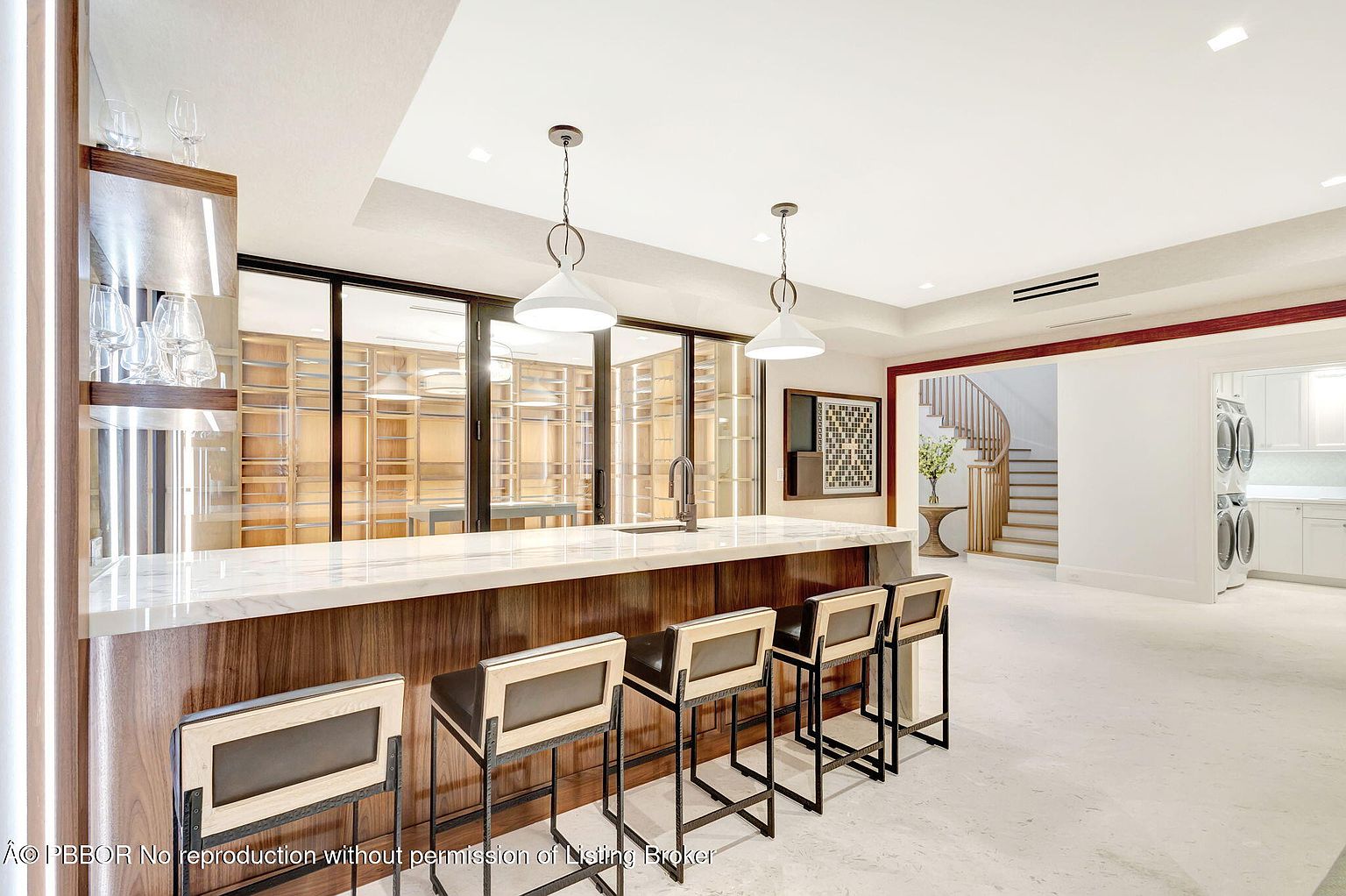 This interior shot showcases a modern kitchen with a stylish bar area. The bar features a marble countertop, dark wood cabinetry, and contemporary bar stools. Behind the bar, there's a wine cellar visible through glass doors, and the open layout leads to a staircase and a laundry area, creating a seamless flow.