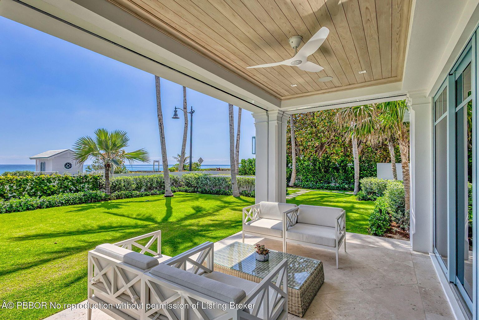 This image showcases a luxurious outdoor patio area, featuring elegant white furniture arranged around a glass-topped coffee table. The patio is covered by a wooden ceiling with a modern ceiling fan, and it offers a view of a lush green lawn, palm trees, and the ocean in the distance. The overall impression is one of sophisticated relaxation and outdoor living.