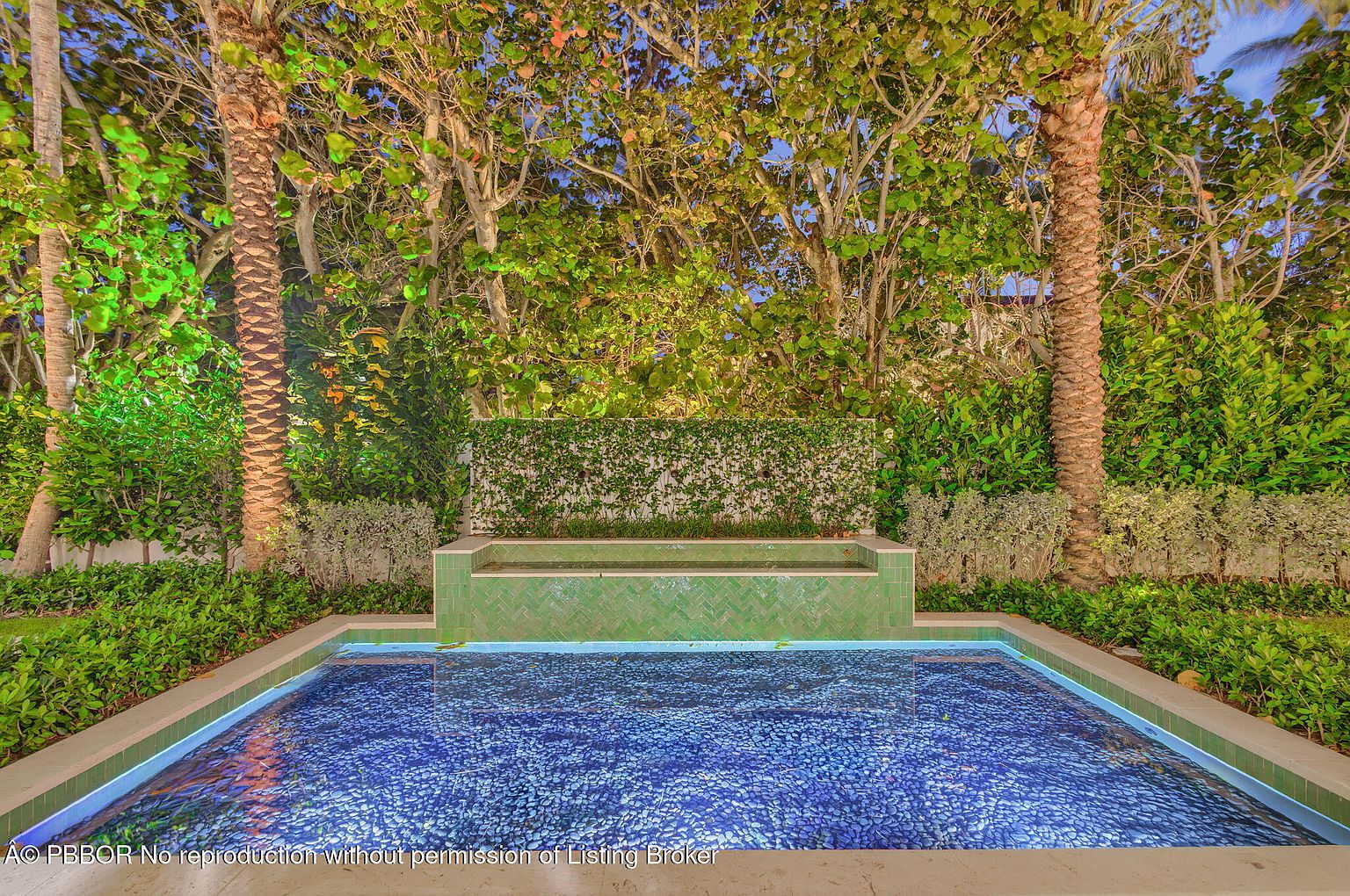 This image showcases a beautifully designed pool area, featuring a rectangular pool with a unique blue pebble bottom. The pool is surrounded by lush greenery, including palm trees and manicured hedges, creating a private and serene oasis. A decorative green tiled water feature adds an elegant touch to the landscape.