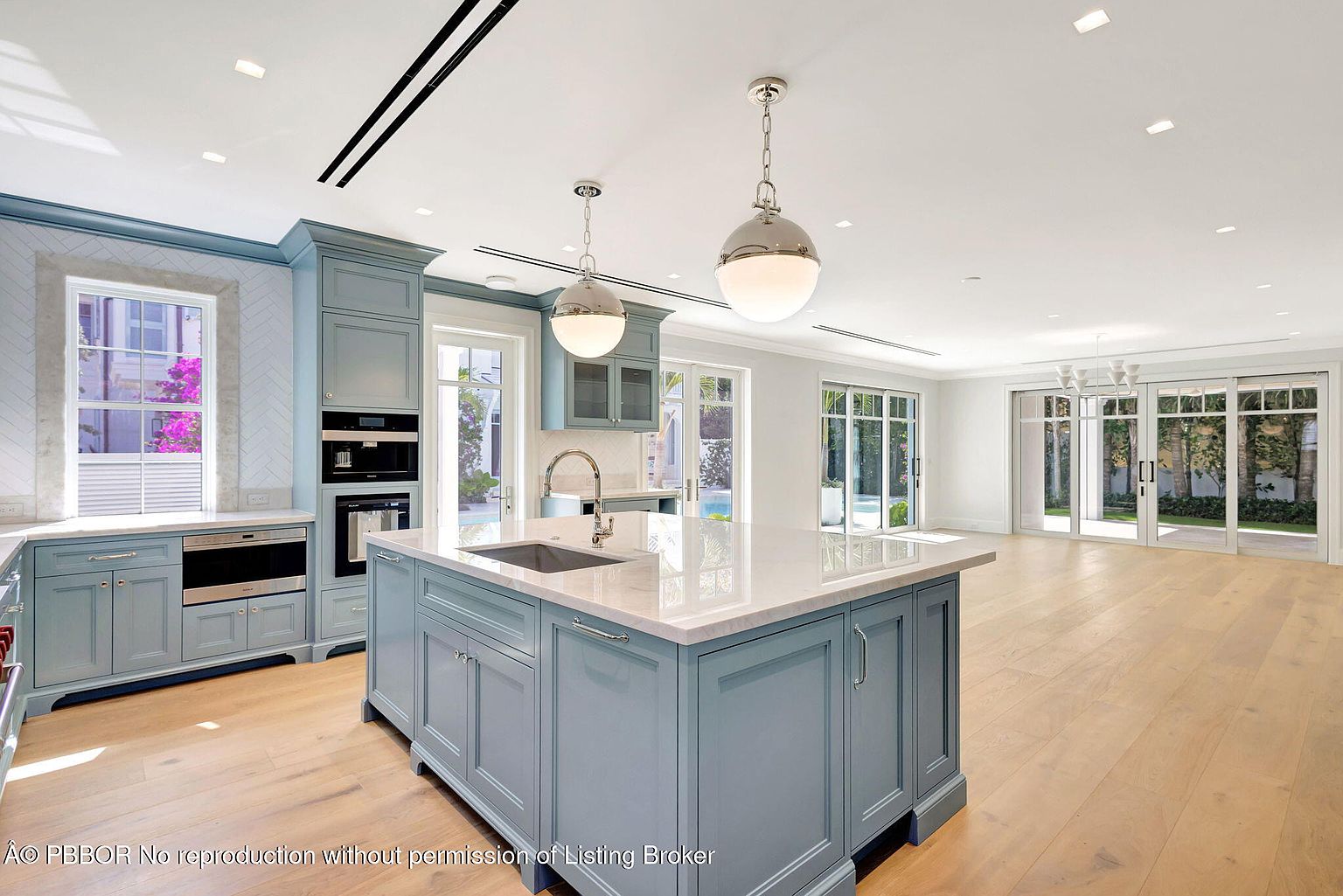 This is a bright and spacious kitchen featuring custom blue cabinetry, a large center island with a white countertop, and stainless steel appliances. The kitchen is well-lit with pendant lighting and recessed lights, and it opens to a large living area with sliding glass doors leading to the outside. The hardwood floors add warmth to the space, creating an inviting atmosphere.