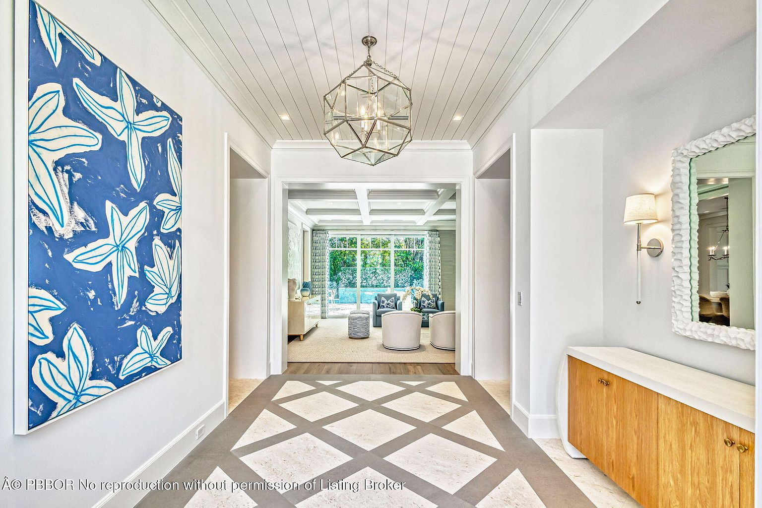 This interior shot showcases a bright and elegant hallway featuring a geometric patterned floor, white walls, and a coffered ceiling with a modern chandelier. A large blue and white abstract painting adds a pop of color, while a glimpse into the adjacent living room reveals a comfortable seating area. The overall impression is one of sophisticated coastal style.