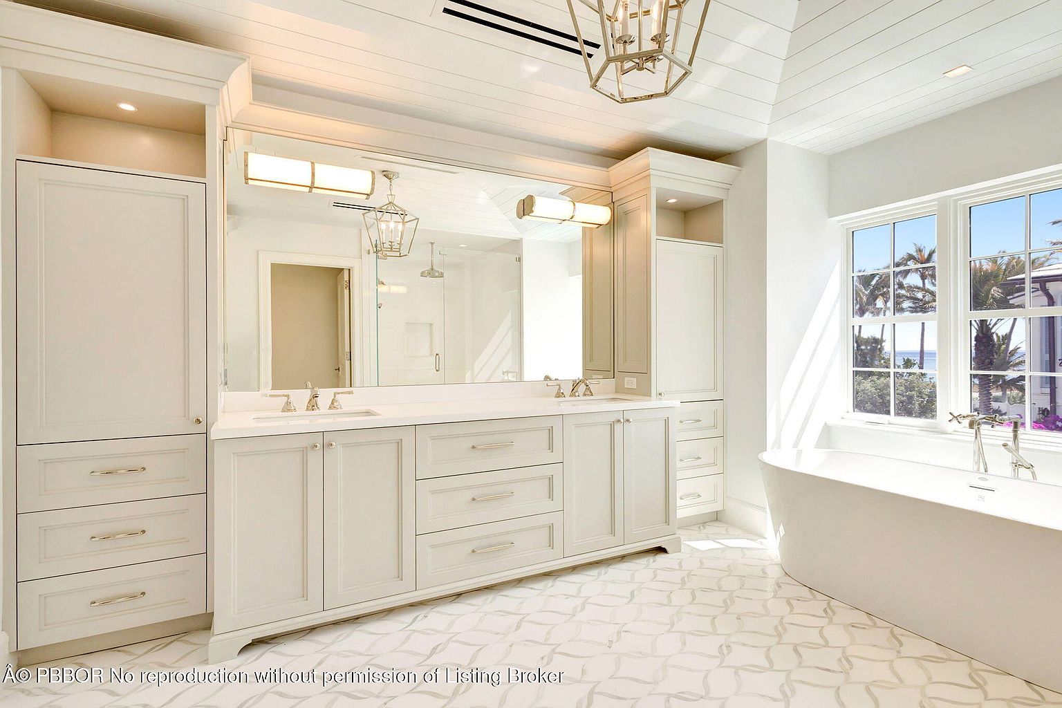 This is a bright and luxurious primary bathroom featuring a double vanity with white cabinetry and a large mirror. The room is well-lit with both natural light from a window overlooking palm trees and stylish overhead fixtures. A freestanding bathtub sits near the window, and the patterned tile flooring adds a touch of elegance.
