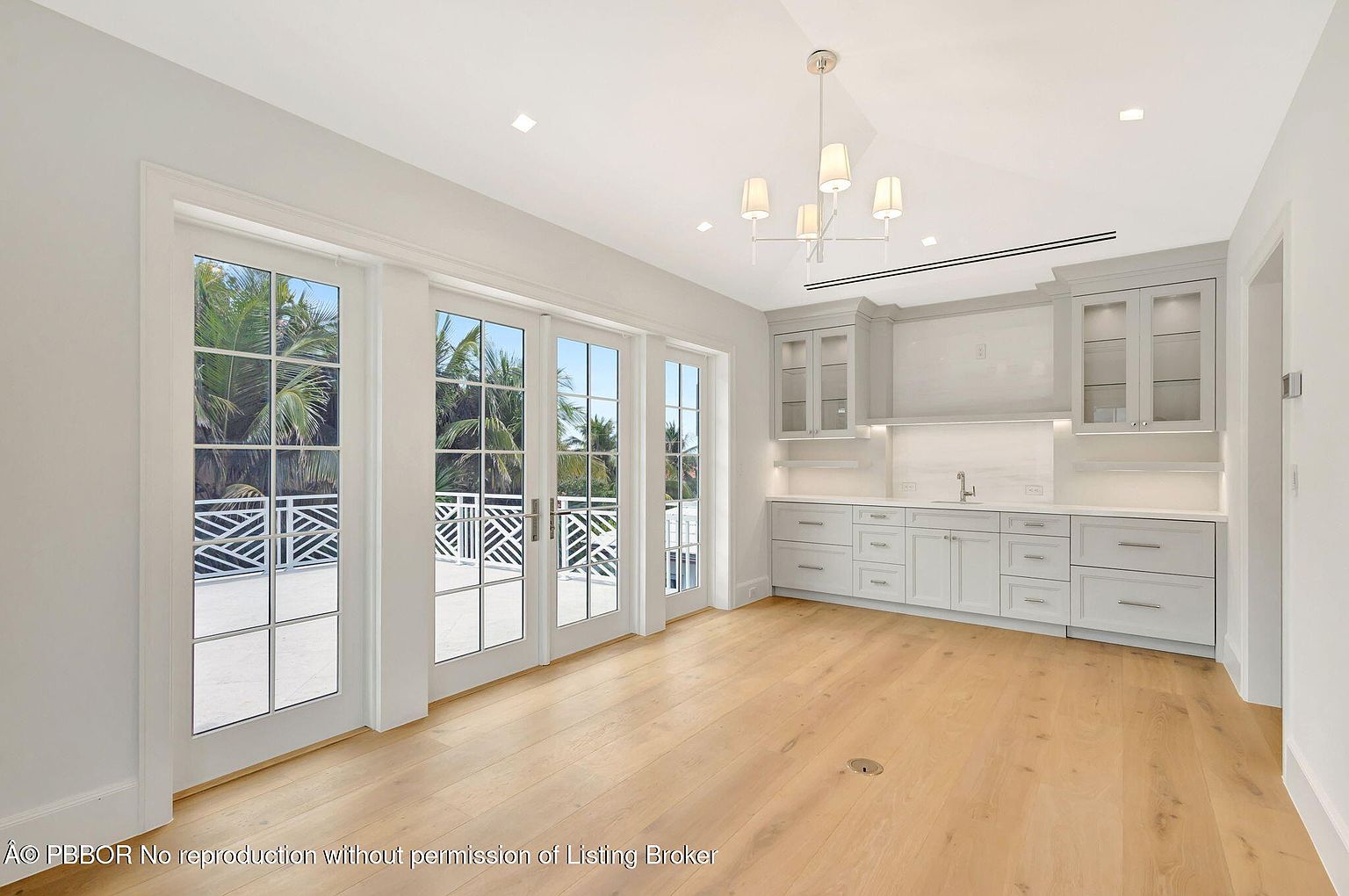 This sunroom features light hardwood flooring and white walls, creating a bright and airy atmosphere. Large glass doors provide access to an outdoor balcony with a decorative railing, offering views of lush greenery. A built-in wet bar with light gray cabinetry and a marble backsplash adds a touch of elegance and functionality to the space.