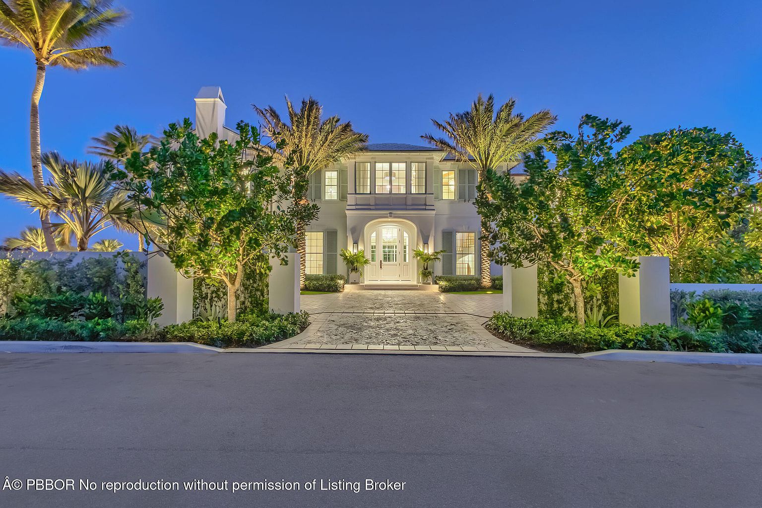 This is a front exterior view of a luxurious two-story home at dusk. The house is painted white with symmetrical design elements, including palm trees flanking the entrance and manicured landscaping. The driveway is paved with decorative stones, leading to a grand entryway with a white door and arched transom window.