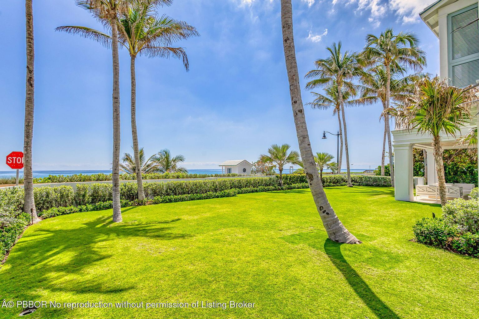 This image showcases a beautifully manicured lawn with palm trees, leading towards a beachfront property. The lush green grass contrasts with the blue sky and ocean, creating a serene and inviting atmosphere. A glimpse of a white building and a stop sign adds a touch of urban elements to the coastal landscape.
