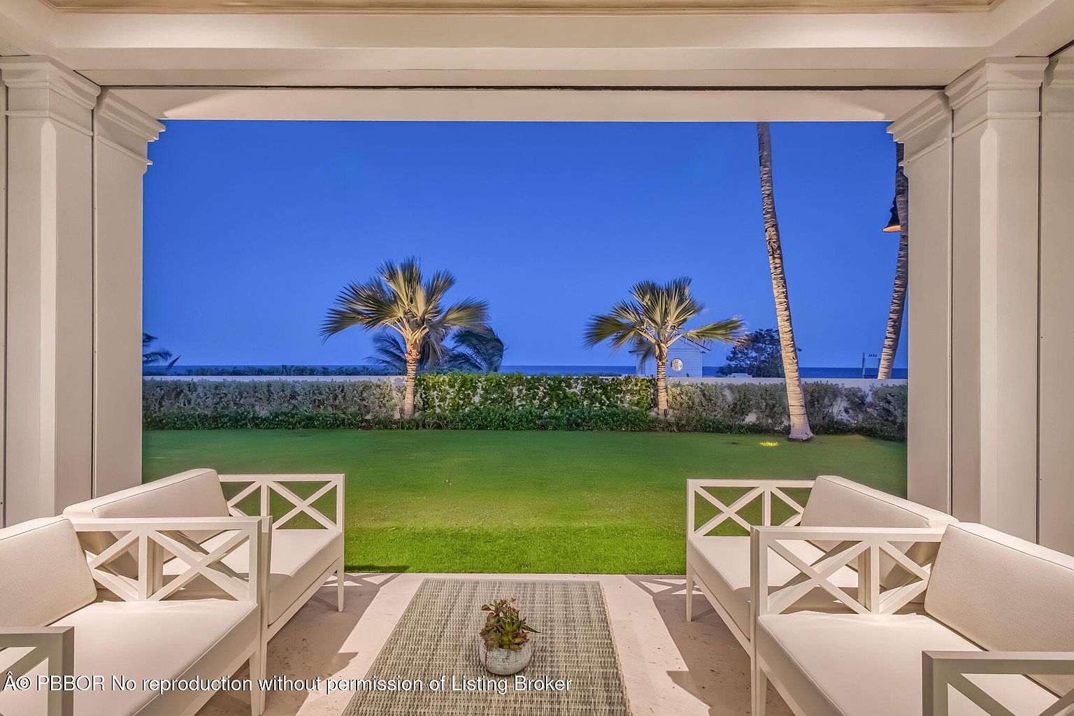 This image showcases a luxurious outdoor patio or balcony area, framed by elegant white columns and architectural details. The space features comfortable seating arrangements, a neutral-toned rug, and a serene view of a well-manicured lawn, palm trees, and the ocean in the background. The overall impression is one of tranquility and upscale outdoor living.