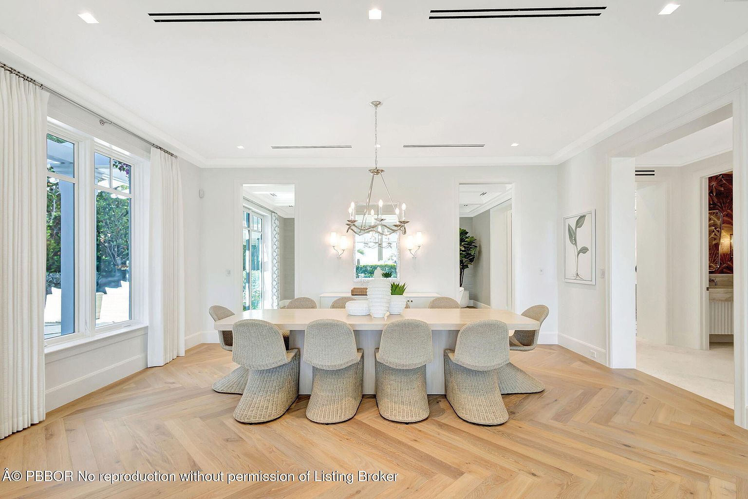 This is a bright and elegant dining room featuring a long, light-colored wooden table surrounded by woven chairs. The room is painted in a crisp white, with large windows providing ample natural light and a view of the outdoors. A chandelier hangs above the table, adding a touch of sophistication to the space, and the herringbone wood flooring adds warmth and texture.