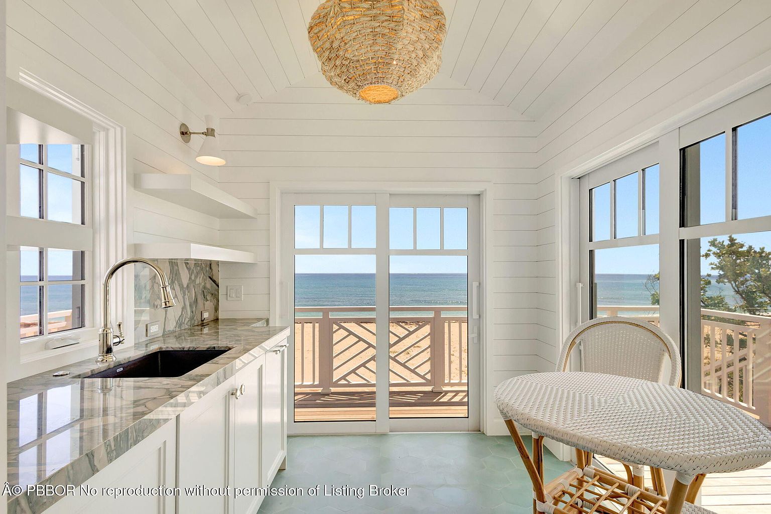 This is a bright and airy kitchen with a coastal aesthetic, featuring white shiplap walls and ceiling, a marble countertop, and a black undermount sink. A woven pendant light hangs from the ceiling, and a sliding glass door leads to a balcony with ocean views. The space includes a small dining area with a round wicker table and chairs, creating a cozy and inviting atmosphere.