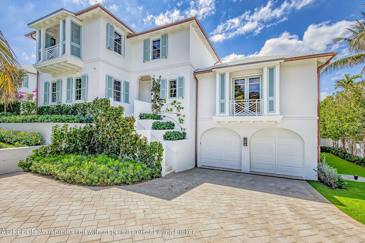 This is a front exterior view of a luxurious two-story house with a white facade and light blue shutters. The property features well-manicured landscaping, including lush greenery and a paved driveway. The architectural style is elegant and inviting, suggesting a high-end residential property.