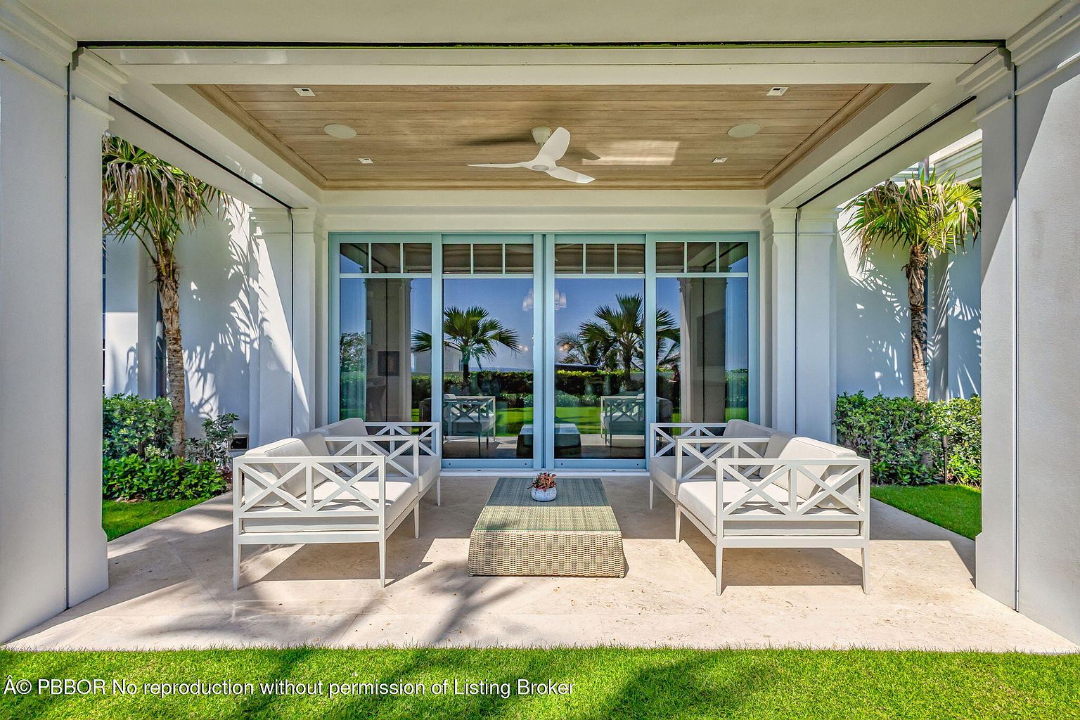 This image showcases a luxurious outdoor patio area, featuring elegant white sofas with a modern design, complemented by a woven coffee table. The patio is covered with a wooden ceiling and a ceiling fan, and the large glass doors offer a seamless transition to the interior, reflecting the surrounding palm trees and greenery. The space is inviting and perfect for relaxation or entertaining.