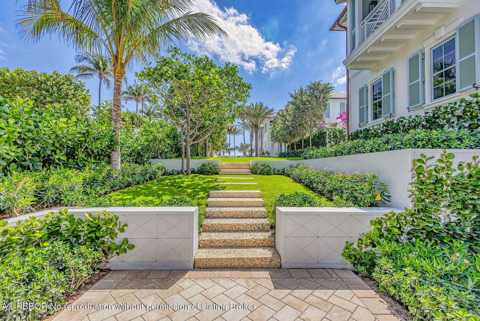 This image showcases a beautifully landscaped yard with a stone staircase leading up a gentle slope. Lush greenery, including manicured bushes and palm trees, surrounds the staircase, creating a serene and inviting atmosphere. The property features a well-maintained lawn and a glimpse of the ocean in the distance, enhancing its appeal.