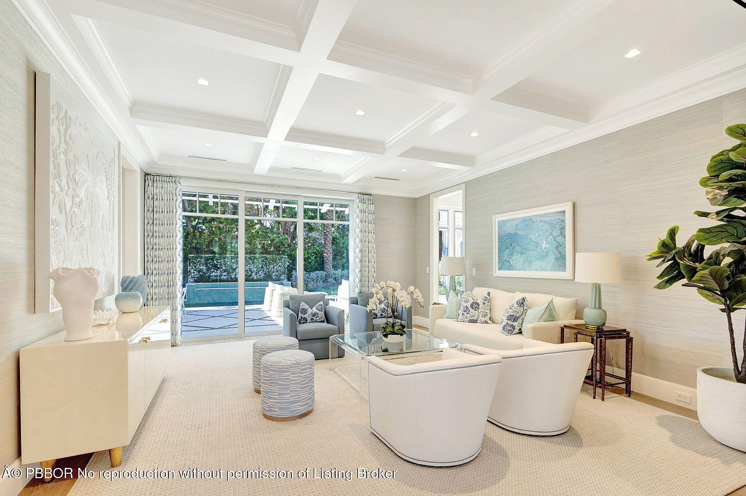 This is a bright and airy living room featuring a coffered ceiling, neutral-toned walls, and plush carpeting. The room is furnished with a white sofa accented with blue and white patterned pillows, two light gray armchairs, and two white armchairs with blue trim. A glass coffee table sits in the center, and large sliding glass doors offer a view of the outdoor greenery and pool area, creating a seamless indoor-outdoor living experience.