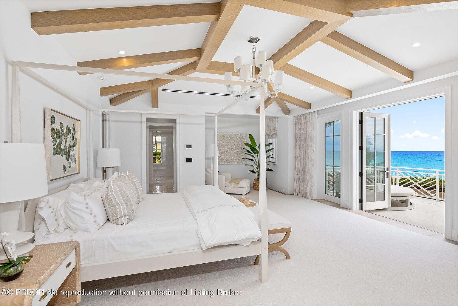 This is a bright and airy primary bedroom featuring a four-poster bed with white linens, complemented by light wood accents in the ceiling beams and bedside table. The room opens onto a balcony with ocean views, enhancing the luxurious and serene atmosphere. The overall design is clean, modern, and coastal-inspired.