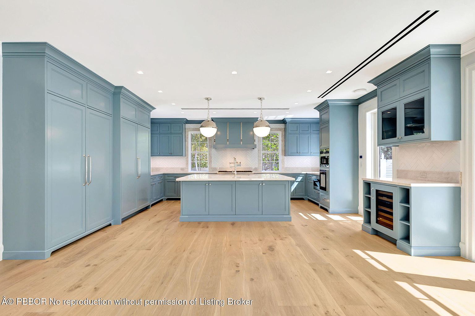 This is a spacious, modern kitchen featuring custom blue cabinetry, stainless steel appliances, and a large central island with a white countertop. The kitchen is well-lit with recessed lighting and pendant lights over the island, and natural light streams in through the windows above the sink. The light wood flooring adds warmth to the space, creating an inviting and luxurious atmosphere.