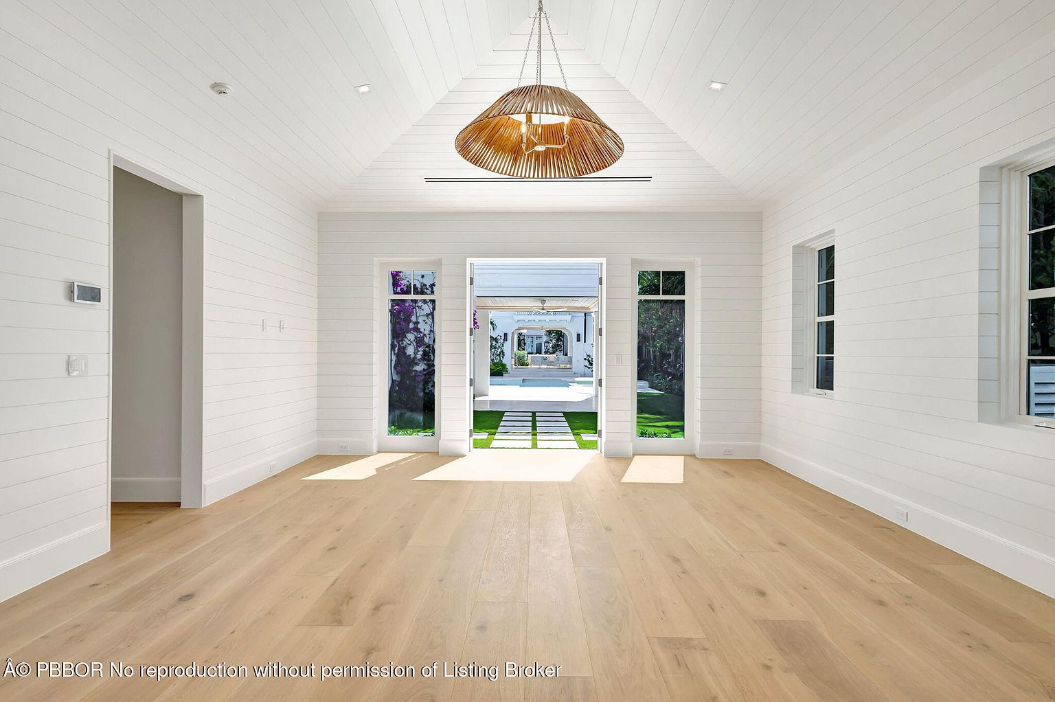 This is an interior shot of a bright and airy living room featuring white shiplap walls and ceiling, light wood flooring, and a large, decorative chandelier. The room opens to an outdoor space through a set of double doors, offering a view of a well-manicured lawn and a covered patio area. The overall impression is one of spaciousness and modern elegance.