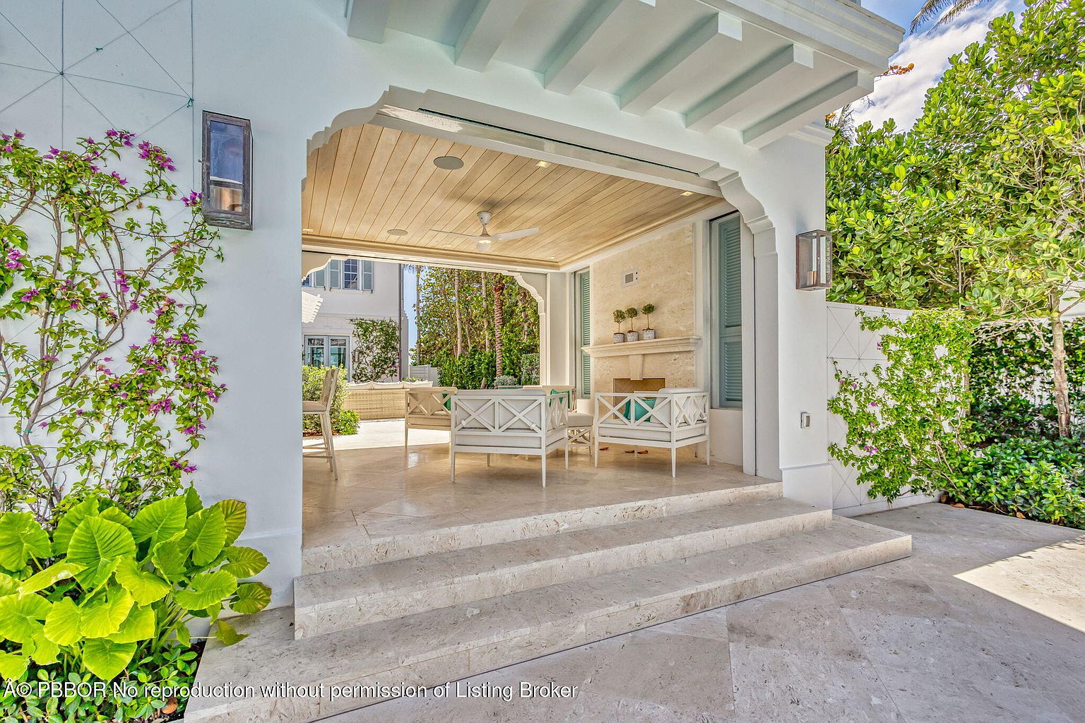 This image showcases a luxurious outdoor patio area, featuring elegant white furniture with blue accents and a cozy fireplace. The patio is accessed by stone steps and is partially covered by a wooden ceiling with a fan. Lush greenery surrounds the space, creating a serene and inviting atmosphere, perfect for relaxation and entertaining.
