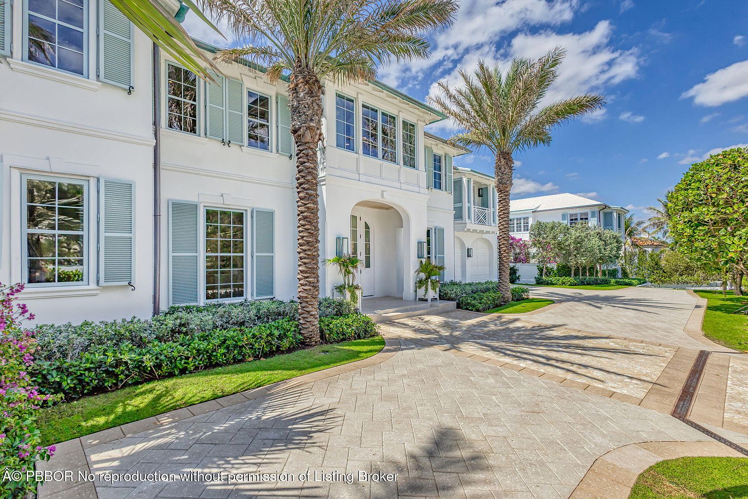 This is a front exterior view of a luxurious two-story white house with light blue shutters and a well-manicured lawn. Two tall palm trees flank the entrance, adding a tropical touch. The driveway is paved with light-colored stones, leading to an arched entryway, and the overall impression is one of elegance and curb appeal.