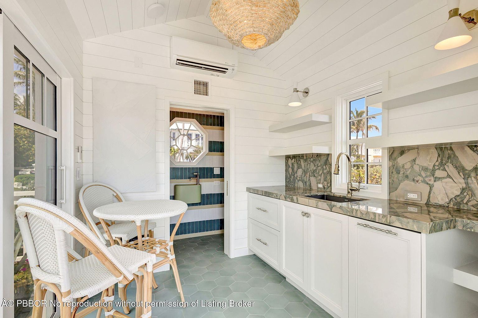 This is a bright and airy kitchen with white horizontal plank walls and a hexagonal tile floor. The kitchen features white cabinets with a marble countertop and backsplash, a stainless steel sink, and a window providing natural light. A small dining area with a round table and wicker chairs is situated near a doorway leading to a bathroom with blue and white striped walls.