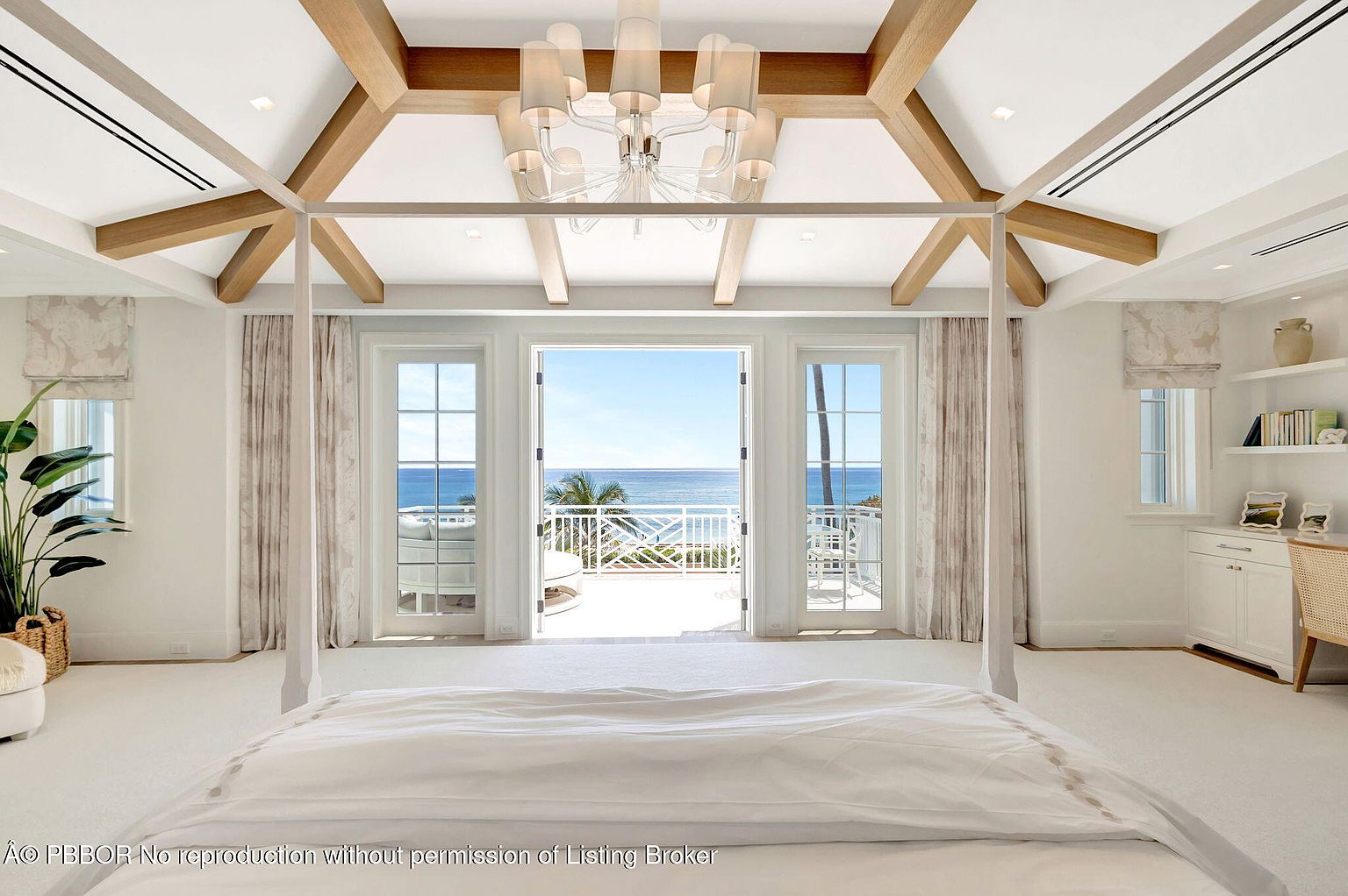 This is a bright and airy primary bedroom with a four-poster bed and a stunning ocean view. The room features a neutral color palette, wooden ceiling beams, and access to a balcony through French doors. The overall impression is luxurious and serene, perfect for relaxation.