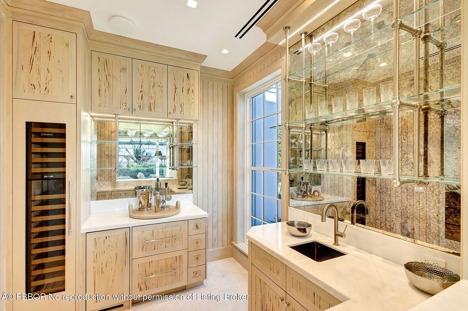 This interior shot showcases a stylish kitchen area with light wood cabinetry and a white countertop. Mirrored backsplashes and glass shelving add a touch of elegance, while a wine cooler is integrated into the cabinetry. The space is well-lit and appears to be designed for both functionality and aesthetic appeal.