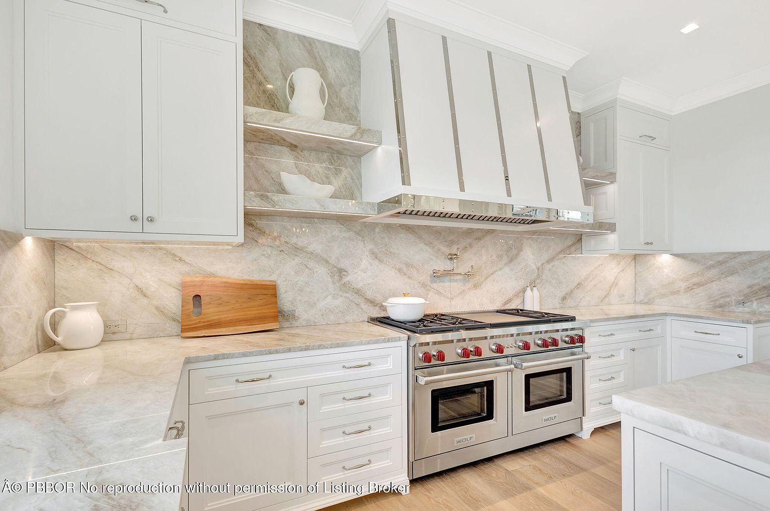 This is a well-lit kitchen featuring white cabinetry, marble countertops and backsplash, and stainless steel appliances. A large Wolf range is the focal point, complemented by a custom range hood. The overall design is elegant and modern, creating a luxurious cooking space.