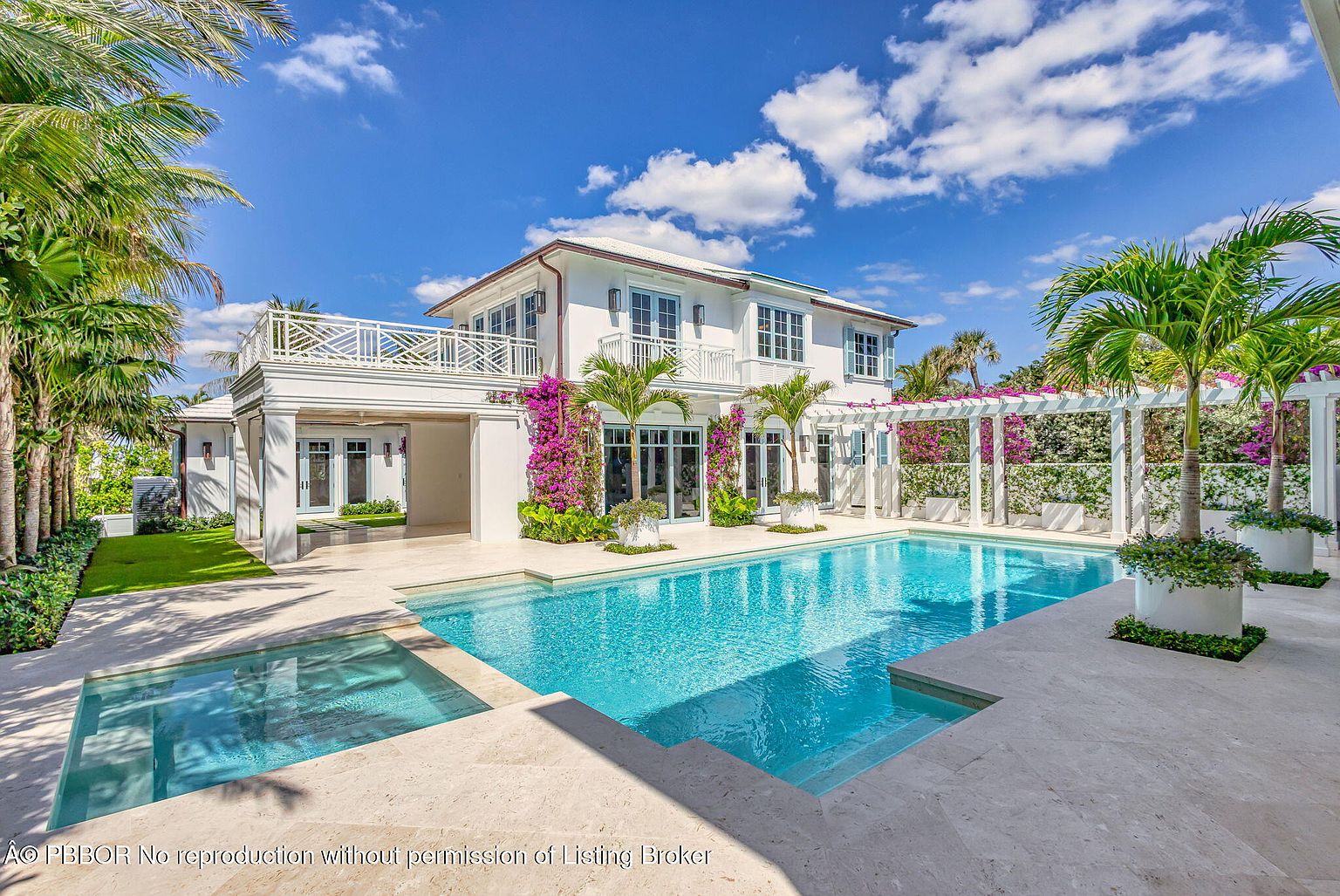 This image showcases a luxurious backyard featuring a pristine pool and spa area. The pool's turquoise water contrasts beautifully with the light-colored stone patio. The property includes a white two-story house with a balcony, surrounded by lush landscaping, including palm trees and flowering vines, creating a serene and upscale outdoor living space.