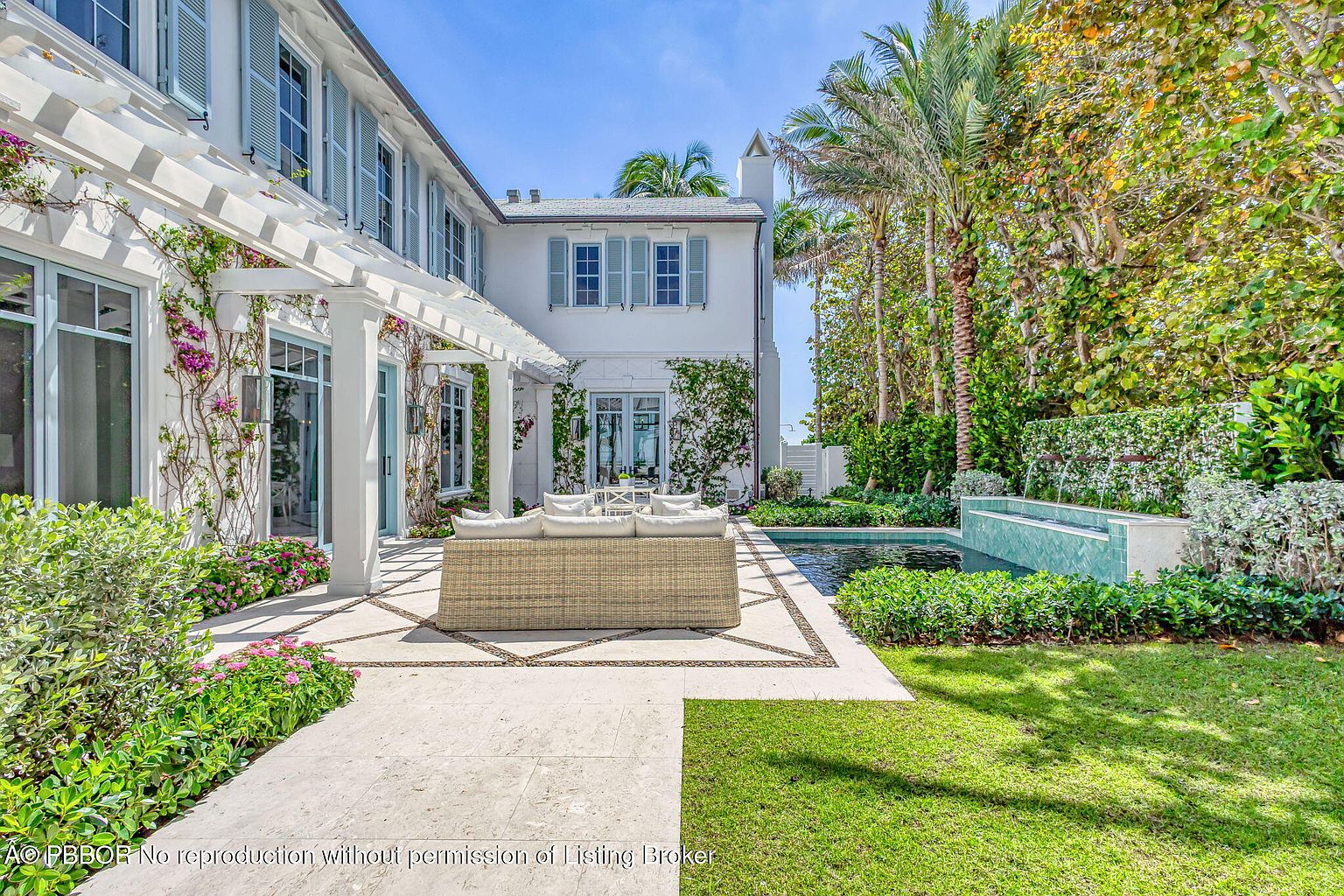 This image showcases a beautifully landscaped backyard featuring a luxurious pool with a modern water feature. A stylish outdoor seating area with a wicker sofa and white cushions is situated on a patterned stone patio. The property is surrounded by lush greenery, including palm trees and manicured hedges, creating a private and serene oasis.