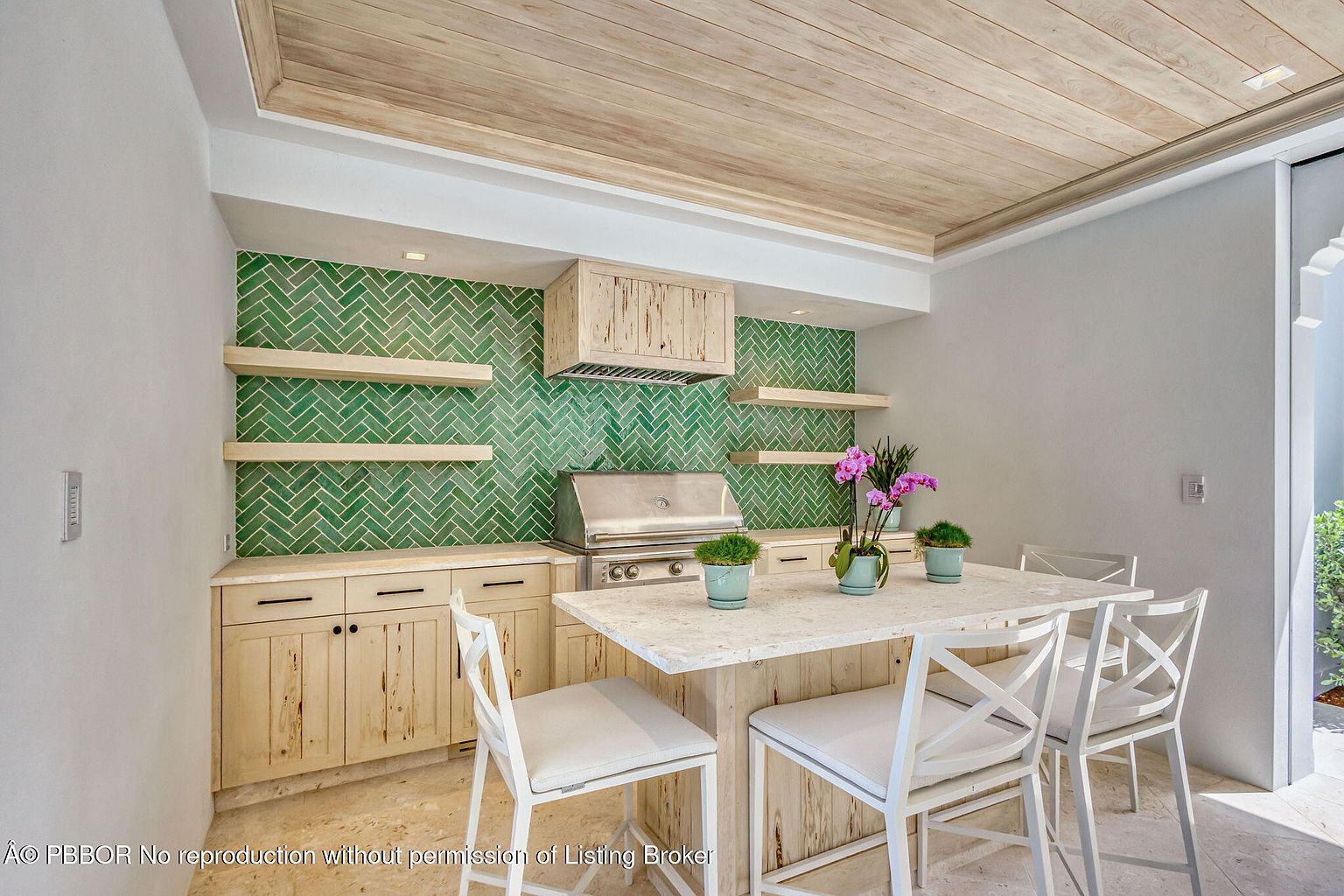 This image showcases a well-appointed outdoor kitchen area with a built-in grill, wooden cabinetry, and a striking green tile backsplash in a herringbone pattern. A central island with bar seating provides a gathering space, complemented by the light-colored stone countertops and the natural wood ceiling. The space is bright and airy, suggesting a luxurious and functional outdoor living area.