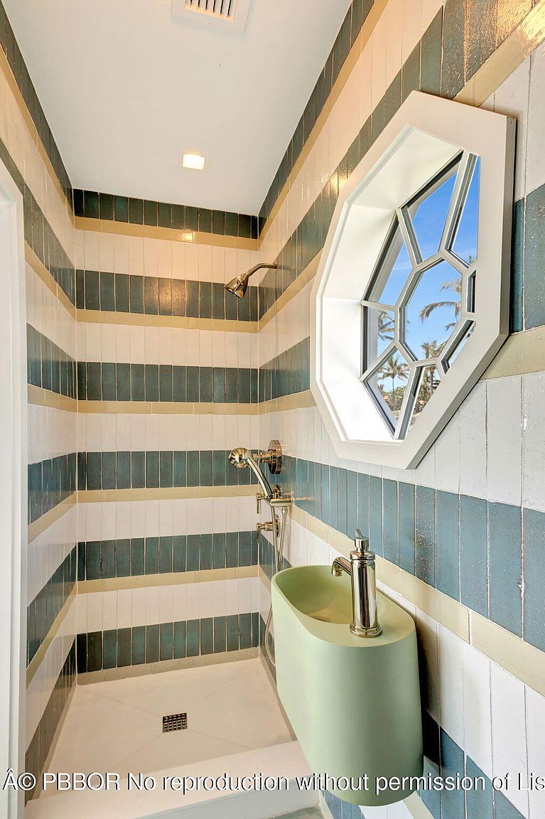 This bathroom features unique striped tile walls in shades of teal, white, and gold, creating a visually striking design. A modern, light green sink is mounted on the wall beneath an octagonal window that offers a view of palm trees. The shower area is compact with a white base and chrome fixtures, contributing to a clean and stylish aesthetic.