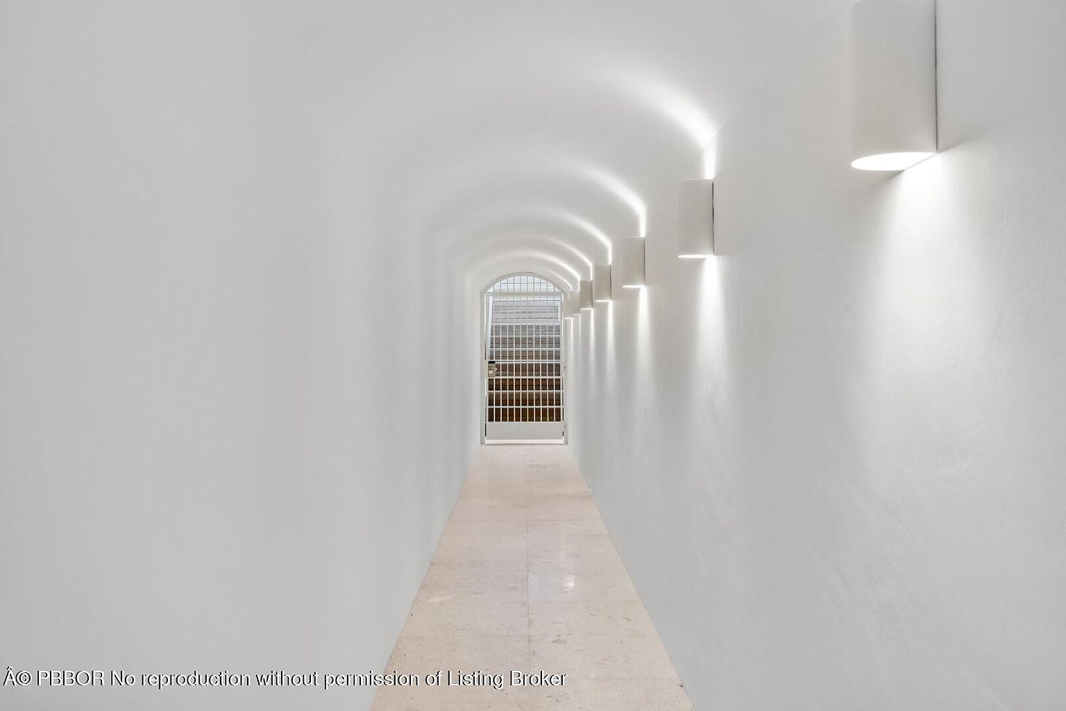 This is an interior shot of a long, arched hallway with white walls and light-colored tile flooring. The hallway is illuminated by modern wall sconces that cast light upwards, creating a dramatic effect. At the end of the hallway is a metal gate, adding a unique architectural detail.