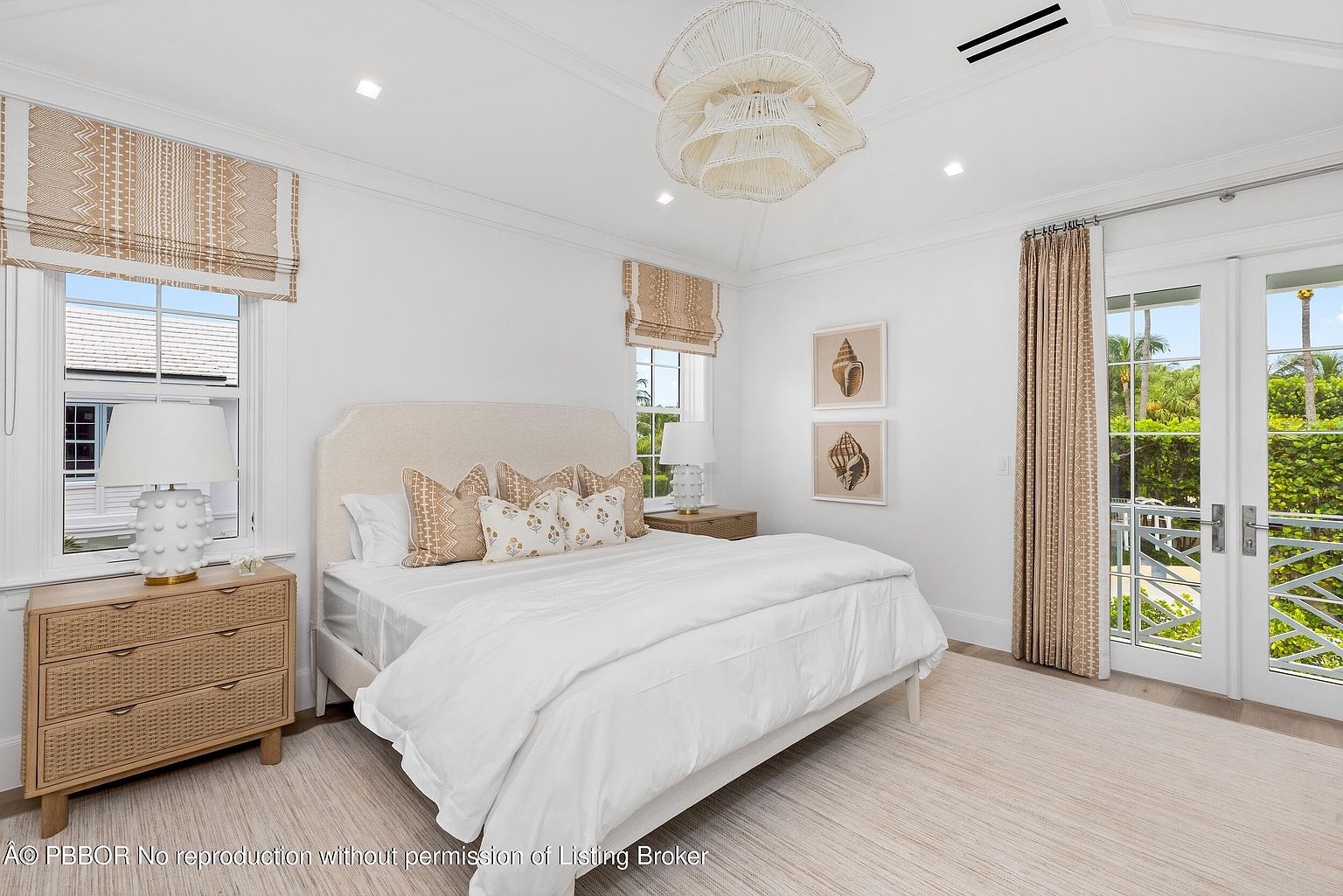 This is a bright and airy primary bedroom featuring a neutral color palette. The room includes a large bed with white linens and decorative pillows, a woven nightstand, and matching window treatments. Natural light floods the space through windows and a glass door, creating a serene and inviting atmosphere.