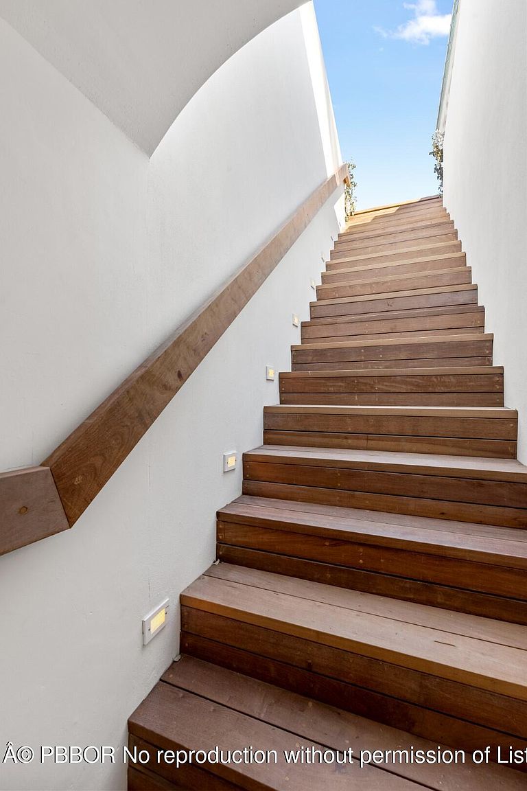 The image showcases a modern staircase with wooden steps and a minimalist design. The staircase is flanked by a white wall with integrated lighting, and a wooden handrail adds a touch of warmth. The upward angle emphasizes the height and length of the staircase, creating a sense of spaciousness and clean architectural lines.