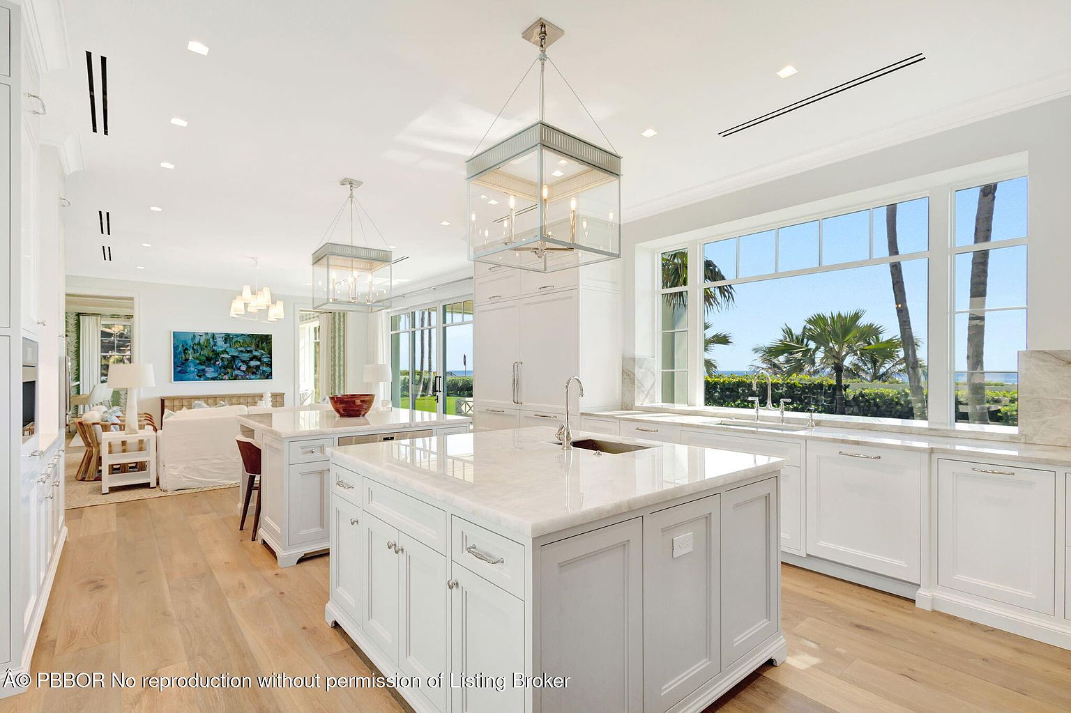 This is a bright and spacious kitchen featuring two large islands with white marble countertops and white cabinetry. The kitchen is well-lit with modern pendant lighting and natural light streaming in from a large window overlooking a tropical landscape. The open floor plan seamlessly connects the kitchen to the living area, creating an inviting and luxurious space.
