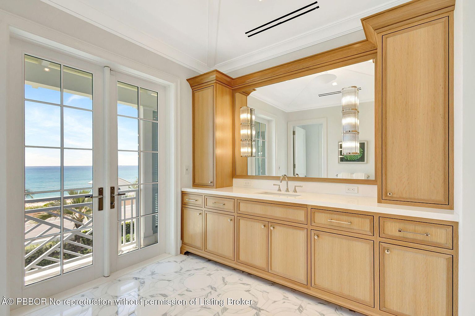 This primary bathroom features a large vanity with light wood cabinetry and a white countertop, complemented by a large mirror and stylish sconces. Double doors open to a balcony with ocean views, enhancing the luxurious and serene atmosphere. The marble flooring adds an elegant touch to the space.