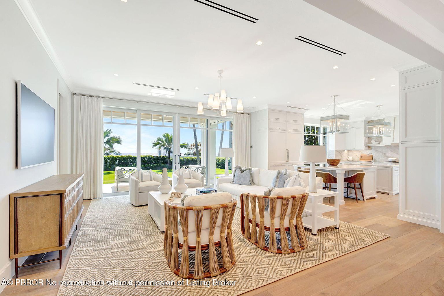 This is a bright and airy living room that seamlessly transitions into a modern kitchen. The living area features comfortable seating, including two unique rope-backed chairs, a white sofa, and additional armchairs visible through the large glass doors that lead to an outdoor area. The kitchen boasts white cabinetry, a marble countertop island with bar seating, and pendant lighting, creating an open and inviting space perfect for entertaining.