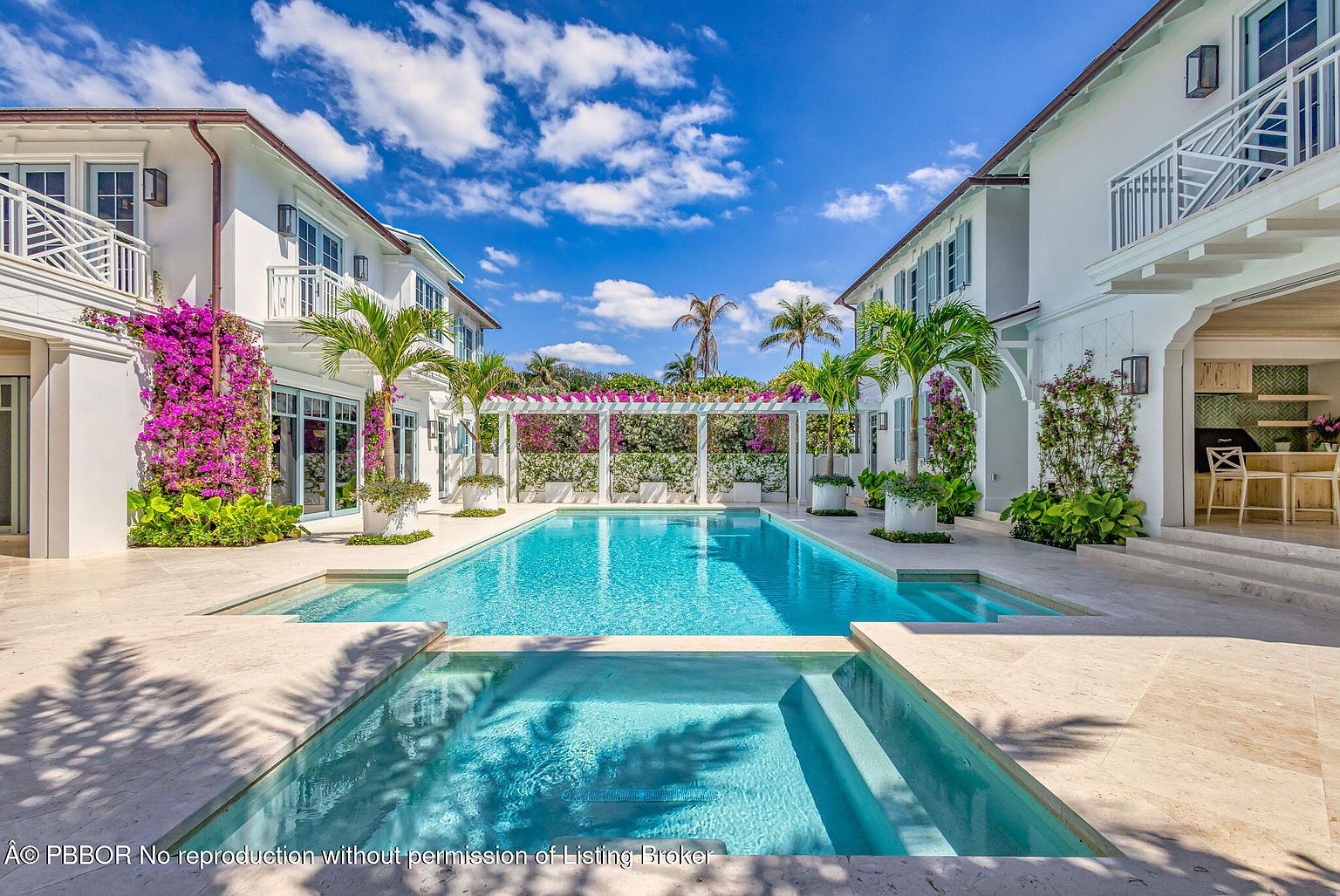 This image showcases a luxurious pool and spa area nestled between two elegant white buildings. The pool features clear turquoise water, surrounded by a light-colored stone patio. Lush greenery and vibrant purple flowers adorn the buildings and a pergola, creating a serene and inviting outdoor space.