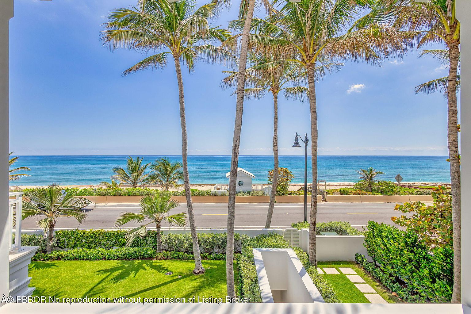 This image showcases a beautifully landscaped yard with lush green grass, palm trees, and manicured hedges. A road separates the property from the beach, offering stunning ocean views. The overall impression is one of tropical elegance and serene coastal living.