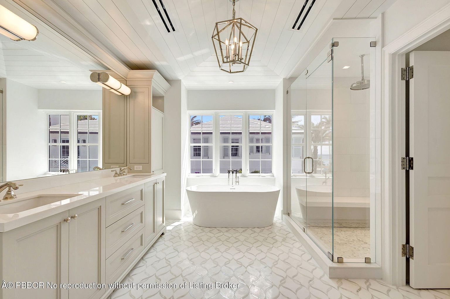 This is a bright and luxurious primary bathroom featuring a freestanding bathtub centered under a window, a glass-enclosed shower, and a long vanity with double sinks. The room is decorated in a clean, white palette with patterned tile flooring and a modern chandelier, creating a spa-like atmosphere. The perspective is a wide shot, showcasing the spaciousness and high-end finishes.
