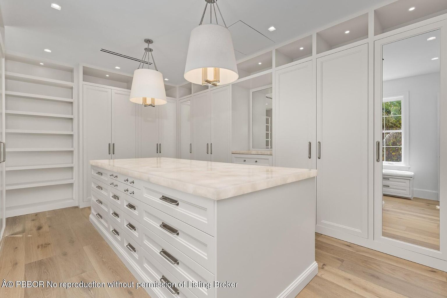 This is a spacious walk-in closet featuring custom white cabinetry, drawers, and shelving. A large center island with a light-colored countertop provides ample storage and workspace. The closet is well-lit with recessed lighting and pendant fixtures, and the light wood flooring adds warmth to the space.