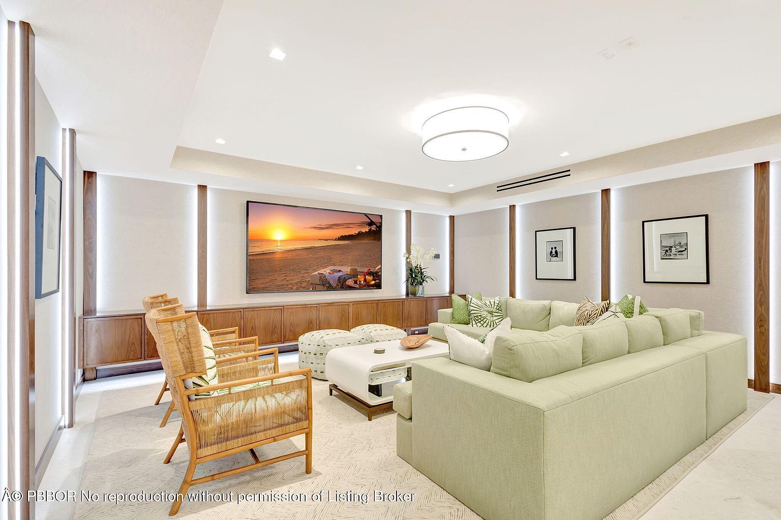 This is a bright and airy living room featuring a large sectional sofa in a light green hue, complemented by rattan chairs and patterned ottomans. A wide-screen television displays a beach scene, mounted above a wooden console. The room is decorated with modern art and recessed lighting, creating a comfortable and stylish space.
