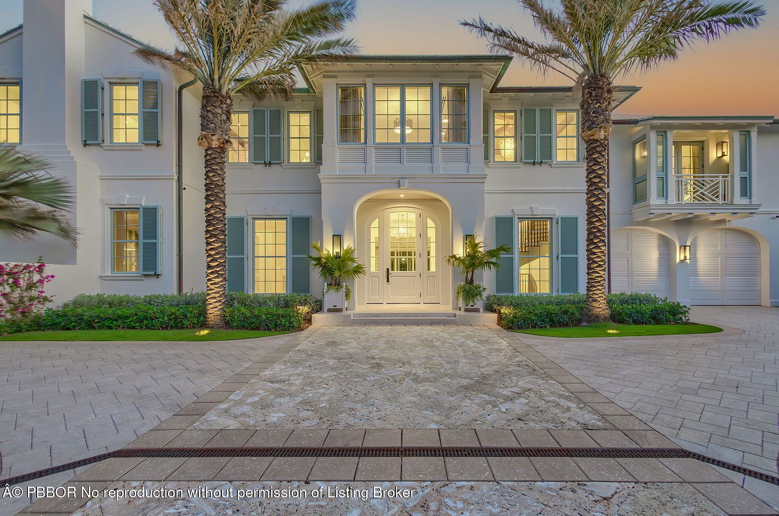 This is a front exterior view of a luxurious two-story home. The house features a symmetrical design with a prominent arched entryway, white facade, and light blue shutters. Palm trees flank the entrance, adding to the tropical feel, while manicured landscaping enhances the curb appeal.
