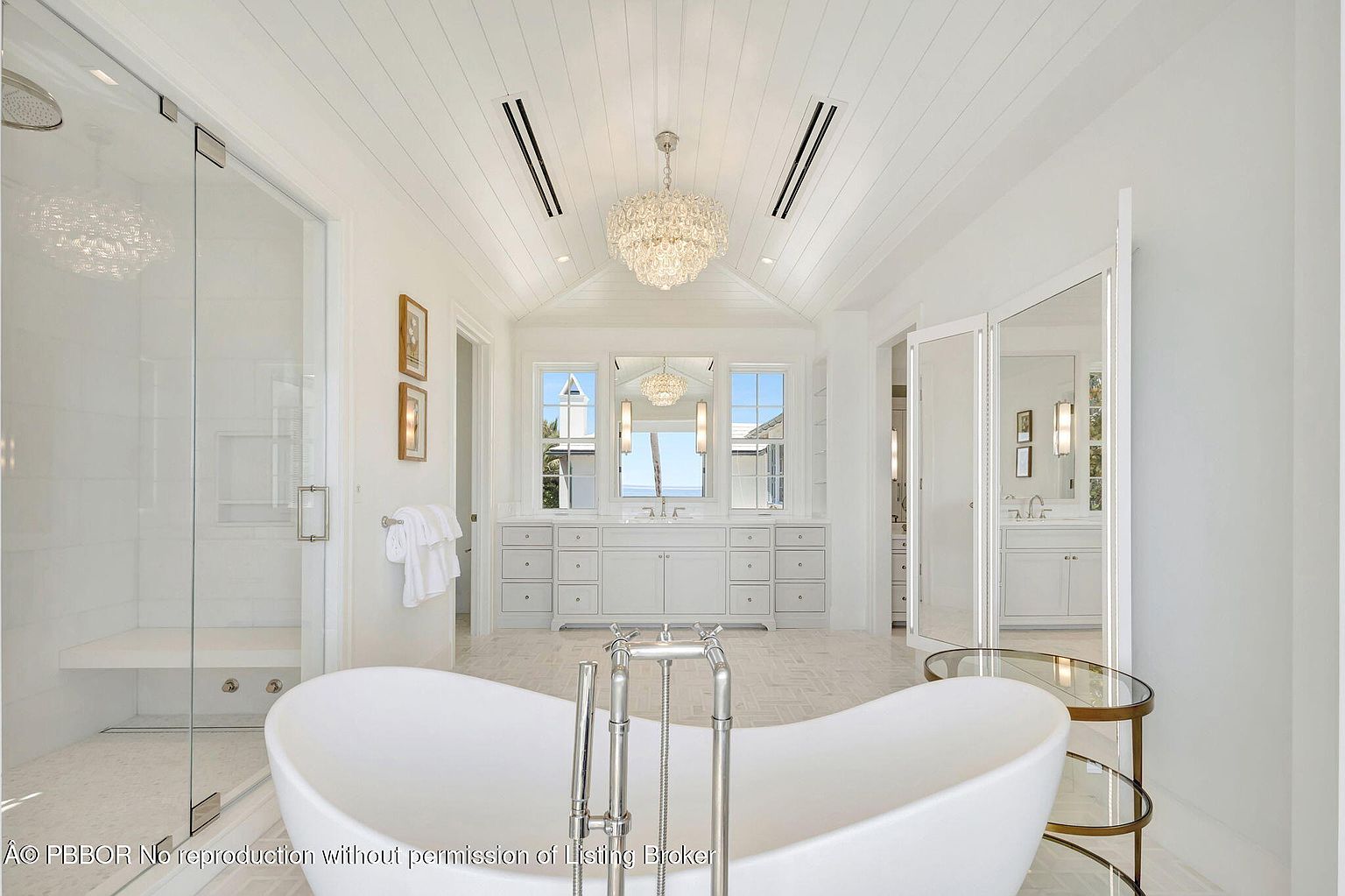 This is a luxurious primary bathroom featuring a freestanding white bathtub as a focal point. The room is bright and airy, with white walls, cabinetry, and a vaulted ceiling with a decorative chandelier. A glass-enclosed shower is visible to the left, and a double vanity with a large mirror is centered in the background, creating a spa-like atmosphere.