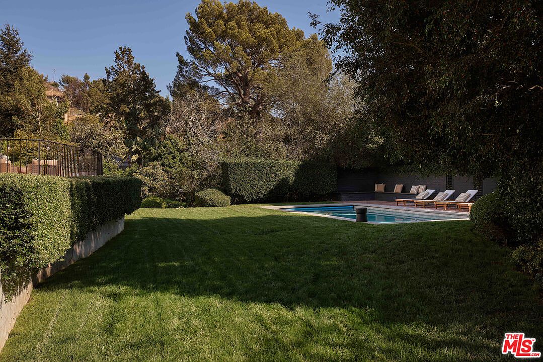 This image showcases a well-manicured backyard featuring a lush green lawn, trimmed hedges, and a swimming pool with lounge chairs. The yard is surrounded by mature trees, providing privacy and a serene atmosphere. The scene evokes a sense of luxury and relaxation, ideal for outdoor entertaining and leisure.