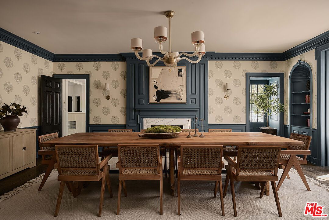 This is a well-appointed dining room featuring a large wooden table with woven chairs, a statement chandelier, and elegant wallpaper with a tree motif. The room is framed by dark blue trim and wainscoting, adding a touch of sophistication. A fireplace with an abstract painting above it serves as a focal point, creating a warm and inviting atmosphere.