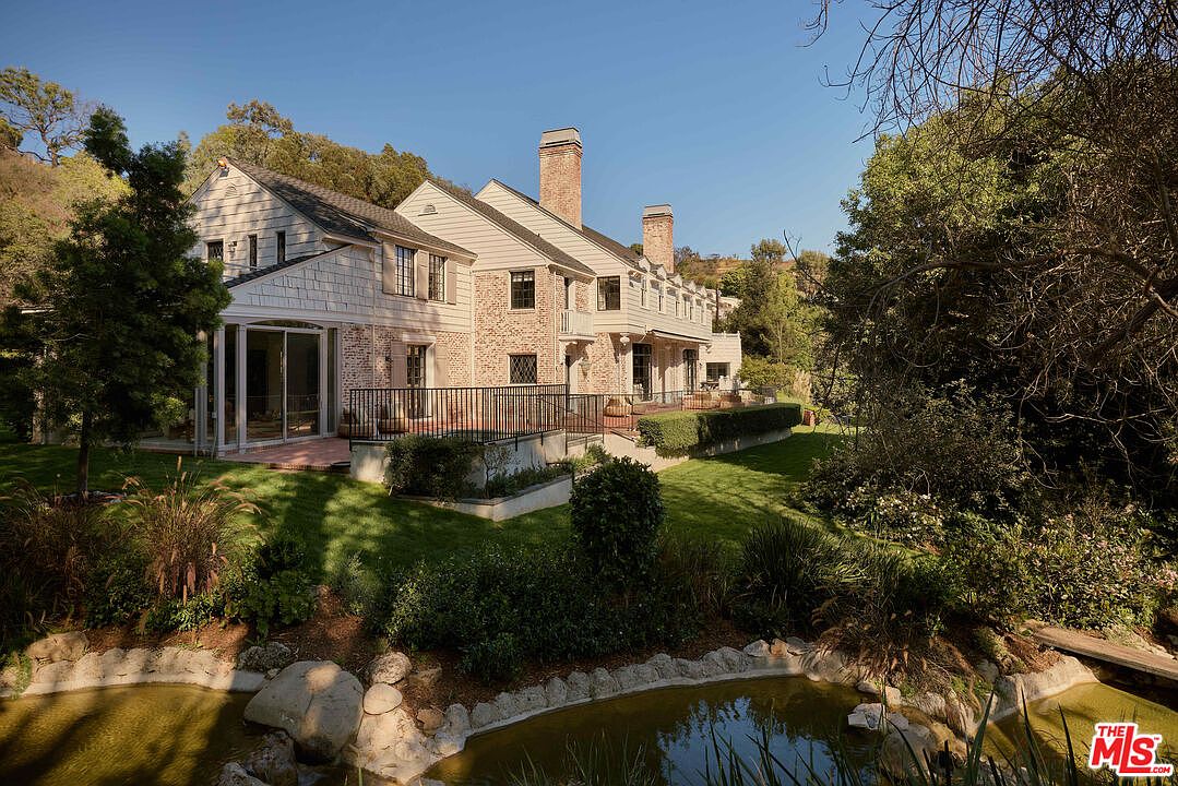 This image showcases the rear exterior of a grand residence, emphasizing its meticulously landscaped yard and garden. A tranquil pond with stone edging and lush vegetation adds a serene touch. The house features a brick facade, multiple chimneys, and a spacious deck area, suggesting a blend of classic elegance and outdoor living.