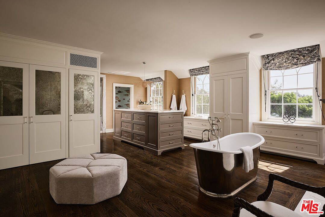 This is a luxurious primary bathroom featuring dark wood flooring, white cabinetry, and a freestanding bathtub. The room includes a central island with drawers, large windows with patterned valances, and mirrored closet doors. The overall impression is elegant and spacious, creating a spa-like atmosphere.
