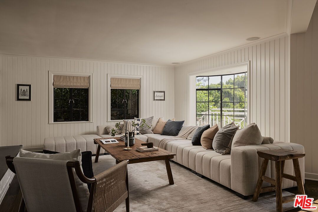 This is a bright and inviting living room featuring white paneled walls and a large sectional sofa with an array of pillows. A rustic wooden coffee table sits in the center of the room, complemented by a woven chair and a wooden stool. Natural light floods the space through multiple windows, creating a warm and comfortable atmosphere.
